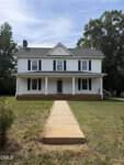 A two-story white house with blue shutters and a covered front porch sits behind a concrete walkway on 218 US 15 in Stovall, surrounded by grass and trees under a partly cloudy sky.