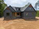 A modern gray house with stone accents at 215 Whistlers Cove, Louisburg, features large front windows and a covered entry. It sits on a dirt lot surrounded by trees, with unfinished landscaping and a curved walkway in progress.