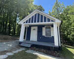 A small blue house with white trim at 217 Lanier Street in Oxford features a welcoming front porch, white door and shutters, and sits beside a gravel walkway and driveway, surrounded by trees and greenery under a clear blue sky.