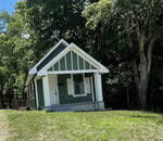 A small green house with white trim sits on a grassy lawn at 219 Lanier Street, Oxford, surrounded by tall trees. The house has a front porch with columns and a steep gabled roof. Sunlight filters through the leaves, casting dappled shadows.