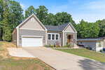 Charming single-story beige house with white trim at 171 Nashua Drive, Louisburg. Features a double garage, sloped driveway, small front porch with steps, minimal landscaping, and is surrounded by green trees under a sunny blue sky.