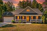 A charming two-story house at 230 N Ridge Drive in Louisburg features a covered front porch, neatly trimmed bushes, and a two-car garage, set against a sunset sky with trees in the background. Warm lights glow from the windows.