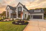 Modern two-story house with large windows, gray and white exterior, attached three-car garage, landscaped front yard, and a clean driveway, set against a sunset sky and surrounded by green grass and trees.