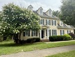 Two-story yellow house with black shutters, three dormer windows, and an entry porch. A large flowering tree and trimmed bushes are in the front yard next to a sidewalk. The sky is partly cloudy.