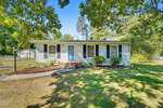 A single-story house at 315 6th Street in Butner features light-colored siding, a blue oval door, black shutters, and several front windows. The yard has green grass, shrubs, a curved walkway, and is shaded by large leafy trees.