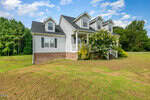 A white, two-story house with a dark roof and brick foundation sits at 1526 Bear Pond Road in Henderson, featuring three dormer windows on a large grassy lawn with trees in the background under a partly cloudy sky.