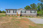 Single-story gray manufactured home with white trim and black shutters at 1305 Schloss Road, Louisburg. Features a small front porch, brick foundation, spacious lot with a gravel driveway, sparse grass, and is surrounded by trees.
