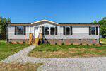 Nestled at 1295 Schloss Road in Louisburg, this single-story modular home features gray siding, black shutters, and a brick foundation. Wooden steps lead to the front door, with a gravel path and tree-lined yard under a clear blue sky.