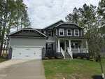 Two-story gray house with white trim at 25 Watermelon Drive, Franklinton. Features a front porch, double garage, stone accents on the lower front, sloped lawn, concrete driveway, and is surrounded by trees.
