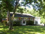 A one-story brick house with a porch and large windows sits at 1025 Carroll Road in Henderson, surrounded by green trees and bushes. The grassy lawn is shaded as sunlight filters through the leaves, and a driveway appears on the right side.
