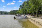 Pontoon boats are docked along the sandy shore at 626 Flemingtown Lane, Henderson, on a calm lake framed by trees and forest under a blue sky. Kayaks and paddleboards rest on the beach for you to enjoy lakeside adventure.
