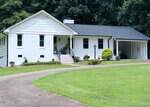 A white single-story house with black roof, front porch, and a carport sits behind a curved driveway at 737 Lakeview Drive in Henderson, surrounded by green lawn and trees. Shrubs and small plants decorate the yard.