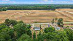 Aerial view of a rural landscape with a few houses surrounded by green trees in the foreground and a large, harvested brown field stretching toward a distant tree line under a partly cloudy sky.