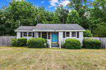 A small, single-story house at 5776 Pearces Road in Louisburg features gray siding, a turquoise front door, white trim, and a fenced yard surrounded by green bushes and trees under a blue sky with scattered clouds.