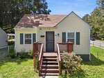 A small, light yellow house at 427 N Clark Street in Henderson, with green shutters and a brown shingle roof, features a wooden front porch with steps and metal door, surrounded by grass, shrubs, and trees on a sunny day.