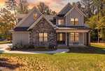 Nestled in Youngsville, 3790 Morning Song Court is a modern two-story house with gray siding, stone accents, black-framed windows, and a covered front porch, surrounded by trees and a grassy yard at sunset.