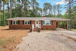 Charming single-story brick house at 4184 Fairport Road, Kittrell, with black shutters, a front porch with steps, two planters, and an attached carport. Surrounded by tall pine trees and a gravel driveway under a blue sky.