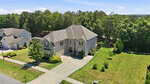 A two-story suburban house with a gray exterior, attached garage, and well-maintained front yard sits beside a spacious green lawn and driveway, bordered by trees and neighboring houses under a clear blue sky.