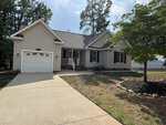Charming single-story beige house at 104 Kiowa Cove, Louisburg, featuring a front porch, attached single-car garage, dark shutters, and a curved driveway surrounded by grass and trees on a sunny day.