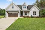 A modern farmhouse-style home at 111 Spring Grove Drive, Oxford, featuring white vertical siding, large windows, a dark double garage door, and a manicured lawn with shrubs beside a tranquil wooded area.