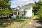 A charming white, single-story house at 604 S Main Street in Louisburg with a covered front porch, several chairs, and surrounded by trees and greenery. The yard features patches of grass and a stone-lined walkway leading to the porch.