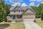 Charming two-story suburban house at 125 Boxwood Drive, Youngsville, featuring beige siding and stone accents, a covered front porch, double garage, manicured lawn, landscaped shrubs, and trees under a blue sky with scattered clouds.