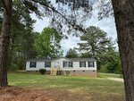 A single-story, light gray manufactured home with white trim and a brick foundation sits on a grassy lot at 141 Abbott Crossings Lane in Henderson, surrounded by tall pine trees under a cloudy sky. Steps lead up to the front door.