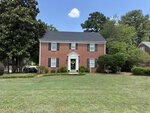A two-story red brick house at 2064 Oxford Road features black shutters, white trim, and a centered front door with a yellow wreath. The Henderson home is surrounded by green bushes and trees, with a spacious, neatly mowed lawn in front.