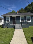 A small, dark green house with a gray roof sits at 124 Sycamore Street in Oxford. It features two front windows, a black door, and a porch with railing. A concrete walkway leads to the steps, surrounded by grass and a trash bin.