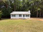 A small, single-story white house with a gray roof sits at 128 Mill Creek Drive in Youngsville, nestled before a wooded area. The front porch has steps and railing, and the grassy yard features a dirt path to the right.