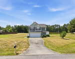 A two-story gray house with white trim and a double garage sits on a large, neatly mowed lawn at 2057 Hidden Pond Drive in Creedmoor, featuring a curved driveway, surrounded by trees and a lake visible under a blue sky.