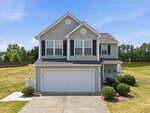 A two-story gray house with white trim, black shutters, and a double garage sits on a manicured lawn with shrubs. A pond and trees are visible in the background under a clear blue sky.