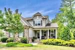 A light gray suburban house at 150 Green Haven Boulevard, Youngsville, features stone accents, a covered front porch with chairs and hanging plants, surrounded by green shrubs, trees, and a well-maintained lawn under a blue sky.