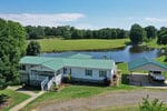A single-story white house with a green metal roof sits beside a pond at 9166 Tilley Road, surrounded by grassy fields and trees. A detached carport with a white truck is nearby under a clear Bullock sky.