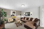 A modern living room at 230 Rosewood Lane in Youngsville features a brown sectional sofa, two beige armchairs, a glass coffee table, houseplants, wall art, a mirror, neutral walls, and a ceiling light.