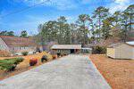 Charming single-story home at 950 Cedar Cove Road, Henderson, featuring a carport at the end of a wide concrete driveway. The yard offers brown grass with shrubs and is framed by tall trees beneath a clear blue sky.