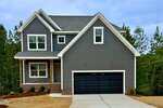A modern two-story house with gray siding, white trim, and a covered front porch at 931 Woodland Road in Creedmoor. The double black garage door faces a concrete driveway and fresh mulch, all set against a scenic backdrop of trees.
