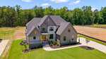 A large modern farmhouse with gray siding, multiple gables, and a well-maintained lawn sits beside a wooded area under a blue sky at 1405 Cottondale Lane in Wake Forest. The driveway curves to the front entryway.