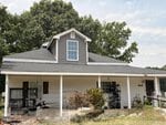 A single-story house with gray siding at 920 Cheves Road in Zebulon, featuring a large front porch, a small dormer window on the roof, and various plants and items on the porch. Trees and blue sky are visible in the background.