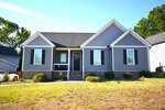 A single-story gray house with white trim and black shutters at 104 Averly Court, featuring a brick foundation, a black front door, and a small porch, set on a grassy yard in the quiet community of Stem with minimal landscaping.