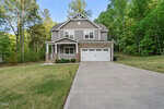 Nestled among green trees at 3440 Lilac Lane in Wake Forest, this two-story suburban house features gray siding, stone accents, a covered front porch, white trim, a double garage, and a large driveway with a well-kept lawn.