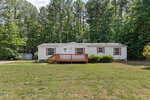 A single-story white house with a wooden front porch sits in a large grassy yard at 1232 Plum Nutty Road, Henderson, surrounded by tall pine trees and dense greenery under a partly cloudy sky.