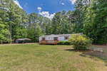 A white manufactured home with a wooden porch sits on a spacious grassy lawn, surrounded by tall trees. A metal carport is visible to the left, and a white bench is on the right near some bushes under a blue sky.