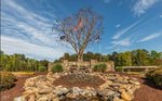 A landscaped area at 125 Red Wing Drive, Louisburg features a stone waterfall, small flower bed, and three flagpoles in front of a large tree, set against a blue sky with wispy clouds and lush greenery.