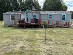 A single-story gray manufactured home with blue shutters at 75 Joe Denton Road in Louisburg, featuring a wooden deck with a table and umbrella, an accessible ramp, and a grassy yard bordered by trees.