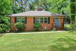 A single-story brick house at 1617 Oakdale Circle with a gray shingle roof, blue front door, and three front windows. Surrounded by green grass, bushes, and trees in Henderson, with a small walkway leading to the entrance.