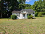 A small white house with a dark roof, front porch, and green shrubs stands on a large grassy lawn at 1222 Serland Mill Road in Henderson. Tall trees and bushes surround the yard under a blue sky with scattered clouds.