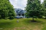A blue two-story house at 20 Tamaran Court, Spring Hope, features dormer windows and a covered front porch, surrounded by green grass and large leafy trees under a partly cloudy sky.