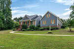 A gray house with yellow shutters at 7101 Evelyn Circle in Zebulon features a front porch with an American flag, neatly trimmed bushes, a curved sidewalk, and a large green lawn surrounded by trees under a blue sky with light clouds.