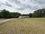 A single-story white house with green shutters sits far back from the road amid a large, open grassy yard at 513 Gay Town Road in Zebulon. A curved driveway leads to the house and an open carport, bordered by trees under a cloudy sky.